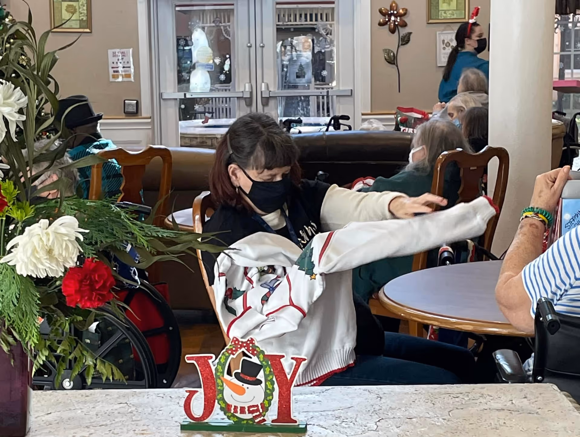 A woman wearing a black face mask is putting on a festive holiday sweater in a common area with other seated elderly people, some in wheelchairs. The room is decorated with holiday-themed items including a 'JOY' sign with a snowman and a floral arrangement with red and white flowers. A staff member wearing a red headband and face mask is visible in the background.