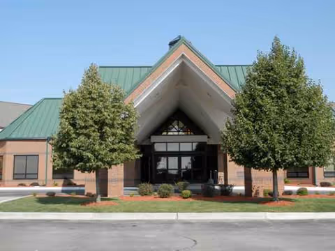Front view of a brick senior living facility entrance with green metal roof and trees.