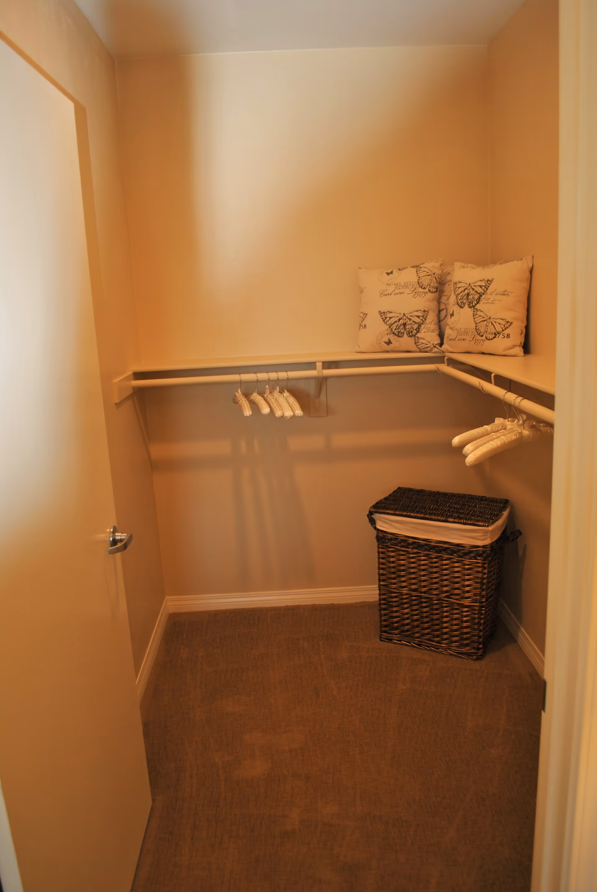 A small walk-in closet with beige walls and carpeted floor. There are white hangers hanging on two sides of the closet rods. Two decorative pillows with butterfly prints are placed on the shelf, and a woven laundry basket with a white liner is on the floor.
