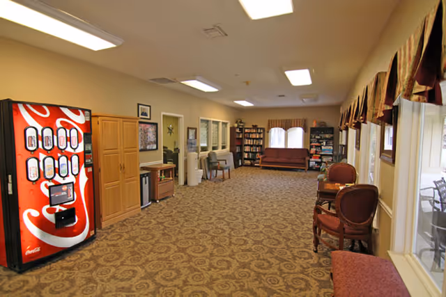 Interior view of a senior living facility common area with patterned carpet, a Coca-Cola vending machine on the left, wooden cabinets, chairs along the right wall beneath windows with valances, and a seating area with a sofa and bookshelves at the far end.
