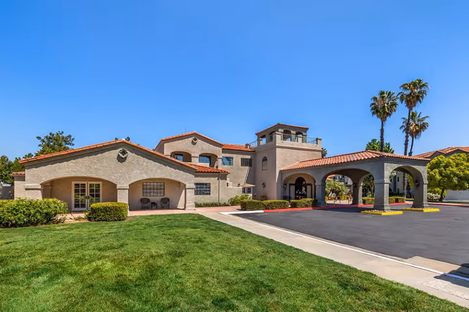 Exterior view of Brookdale Sunwest facility showing a beige stucco building with red tile roof, arched entryway, and a covered driveway. The building is surrounded by green grass, bushes, and palm trees under a clear blue sky.