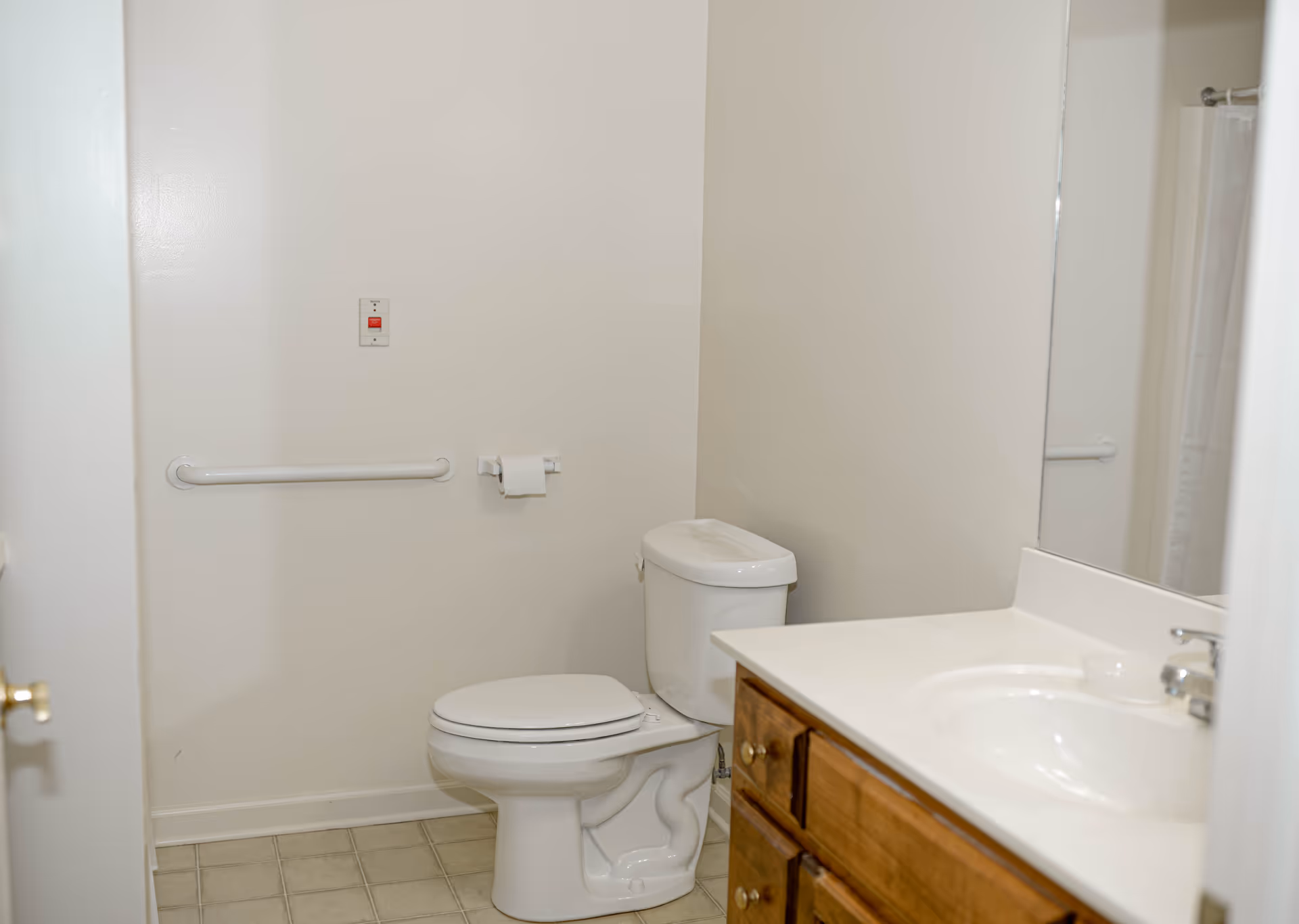 A clean bathroom with a white toilet, a wooden vanity with a white countertop and sink, a large mirror above the sink, a grab bar on the wall, a toilet paper holder with a roll of toilet paper, and a tiled floor.