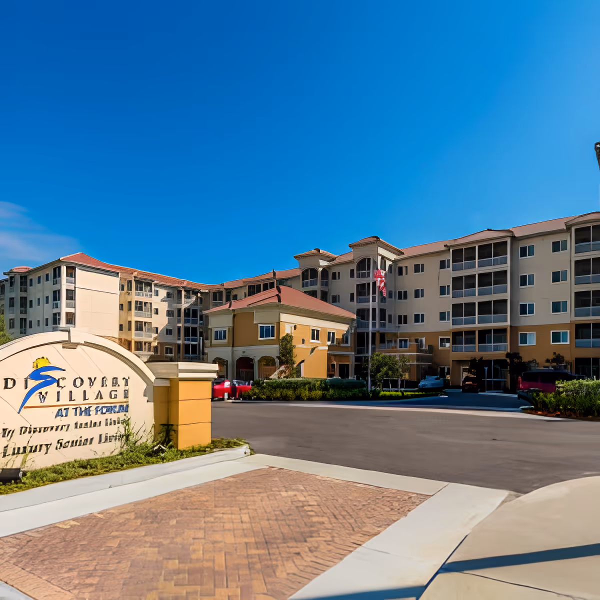 Front exterior of a multi-story senior living building, Discovery Village at The Forum, with a driveway and entrance sign in the foreground.