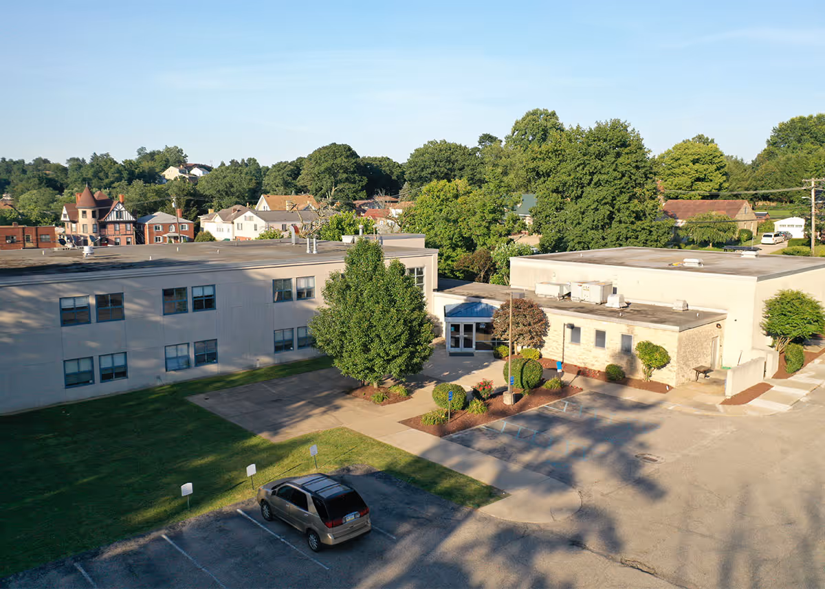 Aerial view of a single-story and two-story building with a parking lot in front, surrounded by trees and residential houses in the background under a clear blue sky.