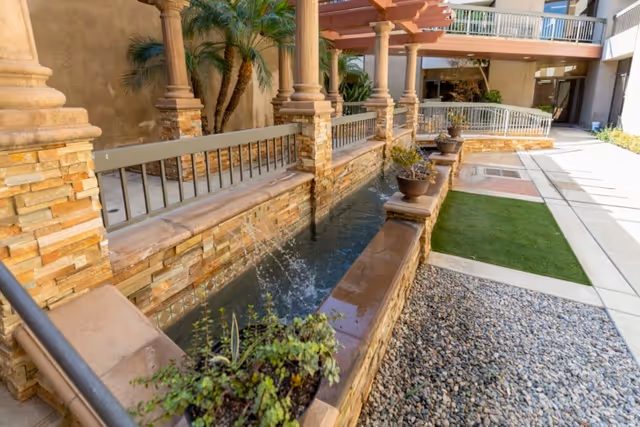Outdoor courtyard with a long stone water feature and small fountains beside columns, planters, and a paved walkway.