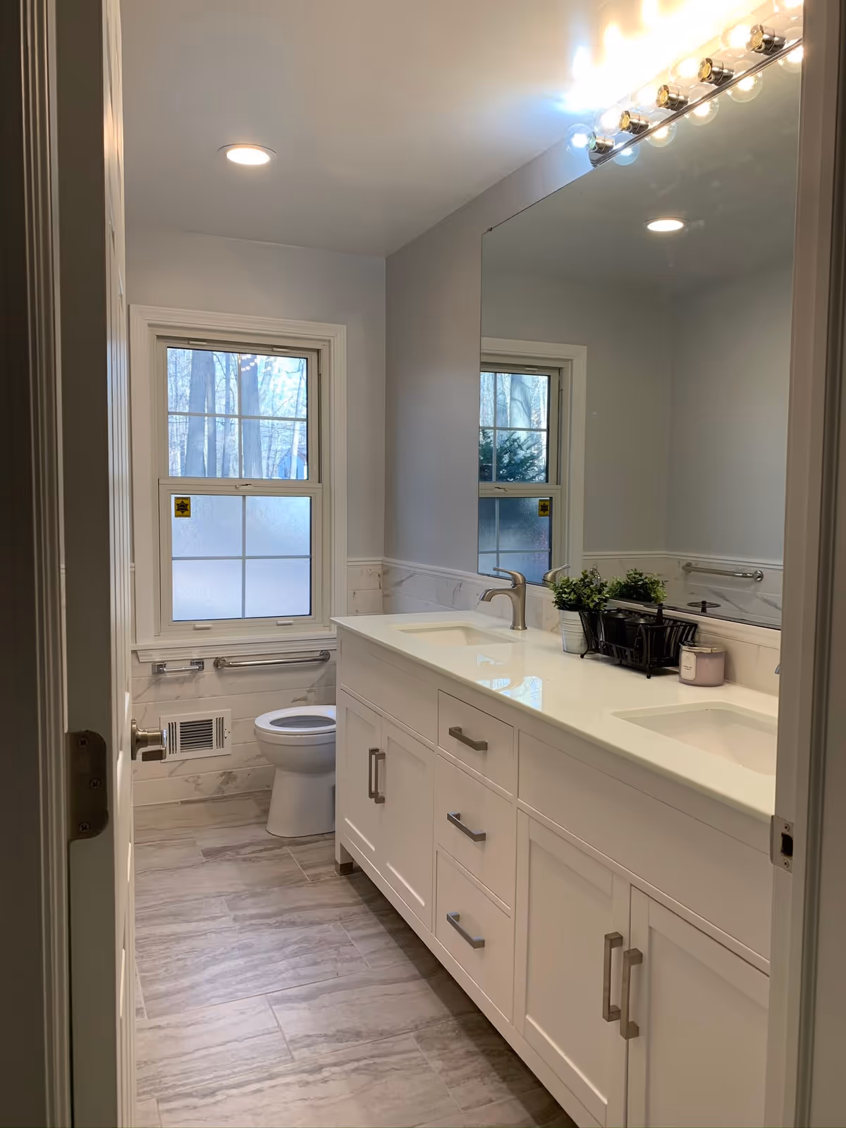 A clean and modern bathroom with a white double-sink vanity, large mirror, toilet, frosted window, and light gray tiled floor and walls.
