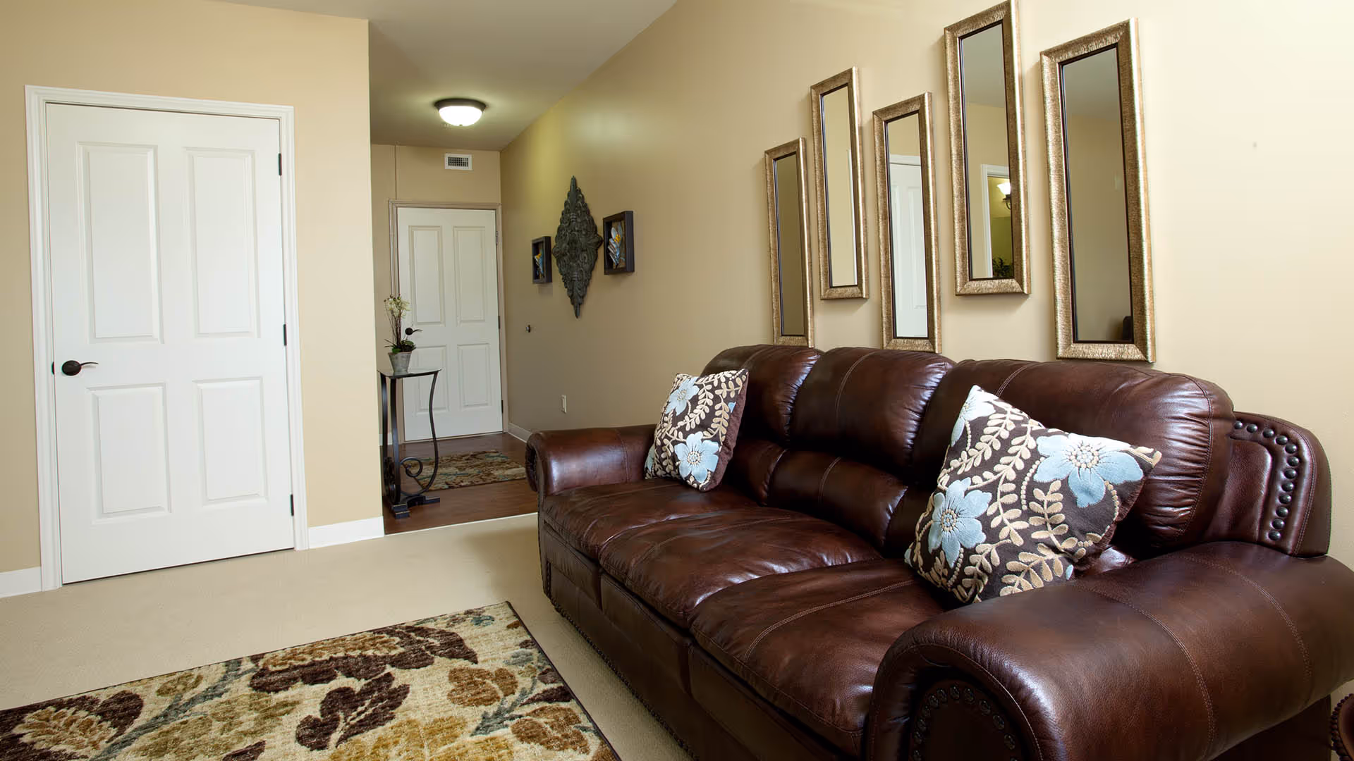 Cozy living room with a brown leather sofa adorned with patterned throw pillows, framed mirrors on the wall, and a view into a hallway and closed doors.