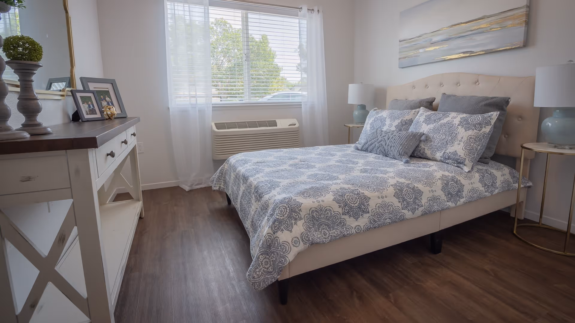 Bright, neatly made bedroom with a patterned blue-and-white bed, matching pillows, nightstands with lamps, a dresser, and a window with blinds.
