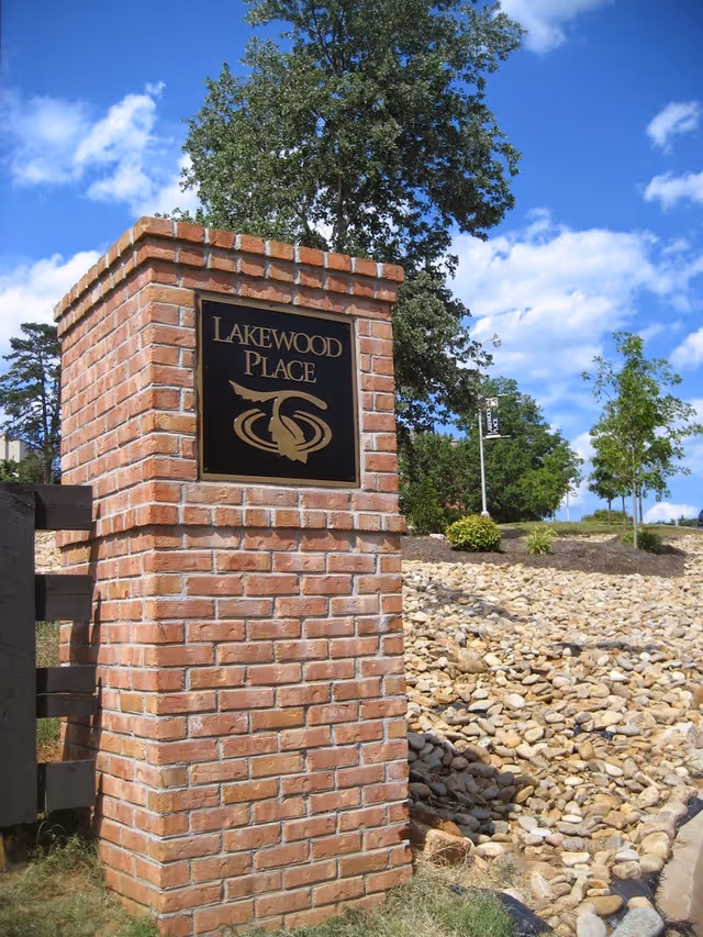 A brick pillar with a black and gold sign that reads 'Lakewood Place' set outdoors with a tree, rocks, and a blue sky with clouds in the background.