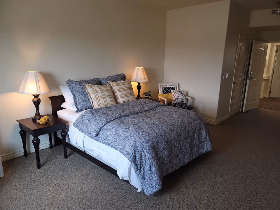 A neatly made bed with blue patterned bedding and multiple pillows, including plaid ones, in a carpeted bedroom. There are two bedside tables with lamps on either side of the bed. A chair with cushions is placed near the bed, and an open door leads to another room or hallway.