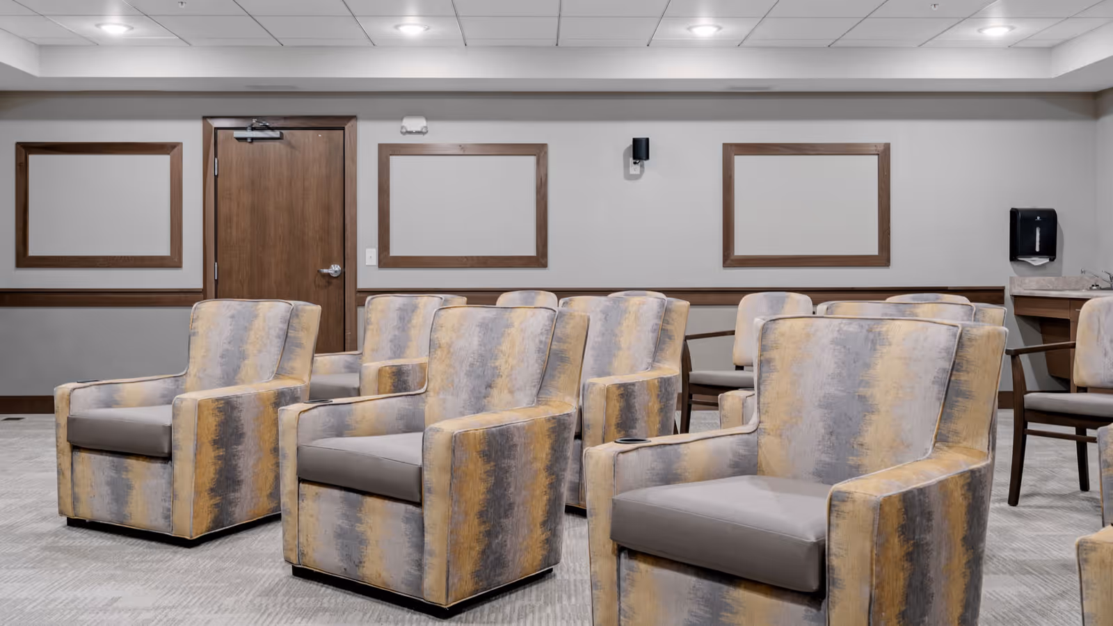 A room with multiple cushioned armchairs arranged in rows facing forward. The chairs have a patterned fabric with shades of gray, yellow, and beige. The walls are light gray with wooden trim and empty wooden frames. There is a wooden door and a wall-mounted black dispenser on the right side.