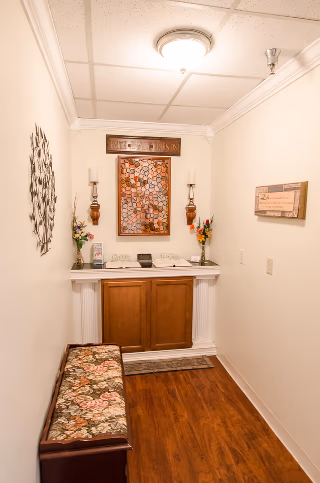 A small, warmly lit hallway with wooden flooring and cream-colored walls. At the end of the hallway is a decorative wooden cabinet with two candle holders and flower vases on top. Above the cabinet hangs a framed mosaic artwork and a sign that reads 'Faith Family Friends'. On the left wall is a metal wall art piece, and on the right wall is a framed inspirational quote. A floral cushioned bench is positioned along the left side of the hallway.