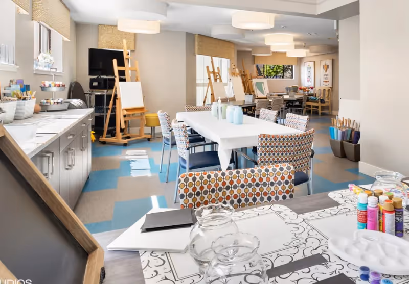 A bright and spacious art activity room with tables covered in white cloths, surrounded by colorful patterned chairs. The room features easels with blank canvases, shelves stocked with art supplies including paint bottles, brushes, and palettes. Large windows allow natural light to fill the space, and modern ceiling lights provide additional illumination.