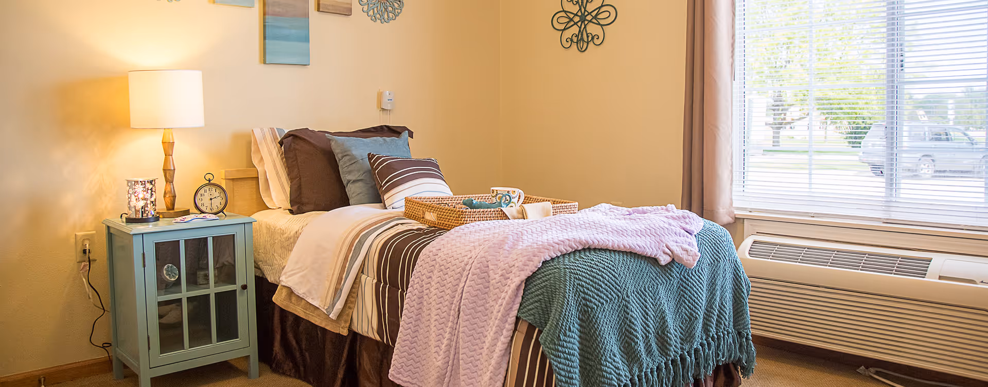 A cozy bedroom in a senior living community featuring a single bed with striped bedding, multiple pillows, and a lavender and teal throw blanket. A wicker tray with a cup and napkin sits on the bed. Next to the bed is a light blue nightstand with a lamp, an alarm clock, and a decorative candle holder. The room has beige walls adorned with wall art and a large window with blinds letting in natural light.