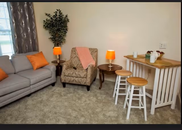 A cozy living room with a gray sofa adorned with an orange pillow, a patterned armchair with a pink throw draped over it, two small wooden side tables each with an orange lamp, a tall green plant in the corner, and a small wooden bar table with two stools. The room has beige carpet and light-colored walls.