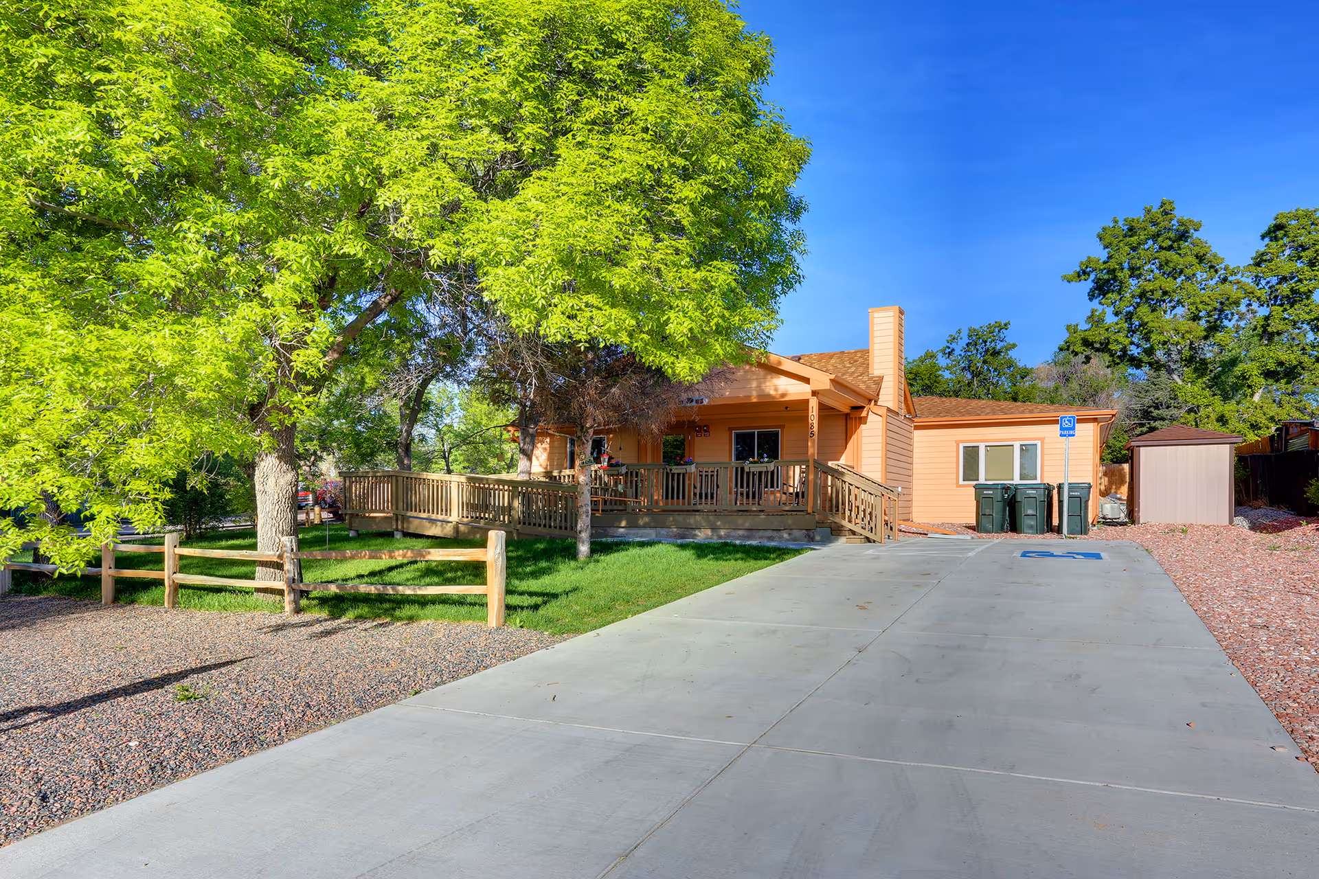 Single-story peach-colored building with a front porch and wheelchair ramp, large green trees, and a long concrete driveway.