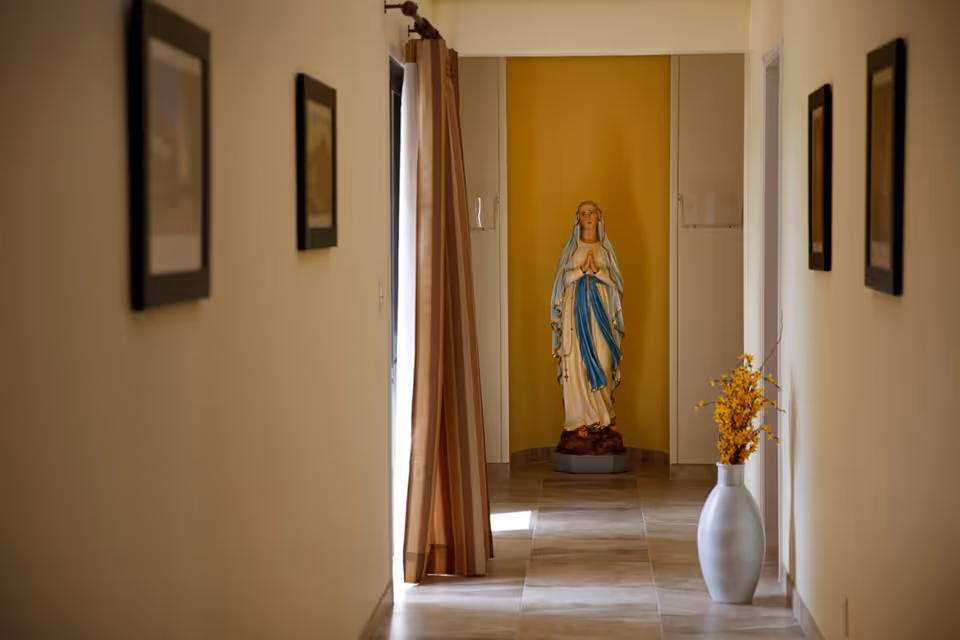 A sunlit interior hallway with framed pictures on the walls, a vase of yellow flowers, and a statue of the Virgin Mary at the far end.