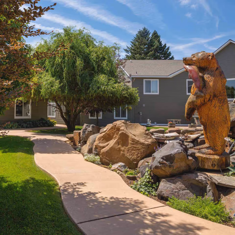 A winding concrete pathway through a landscaped garden area with green grass, trees, and large rocks. To the right, there is a wooden bear statue holding a fish. In the background, there is a gray building with windows under a blue sky with some clouds.