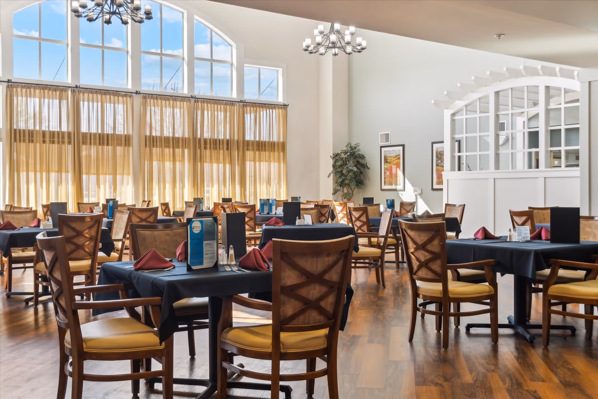 A bright dining room with large arched windows covered by sheer curtains, wooden chairs with yellow cushions, tables set with black tablecloths, red folded napkins, menus, and salt and pepper shakers. The room has wooden flooring, two chandeliers, framed artwork on the walls, and a white partition with glass panels.
