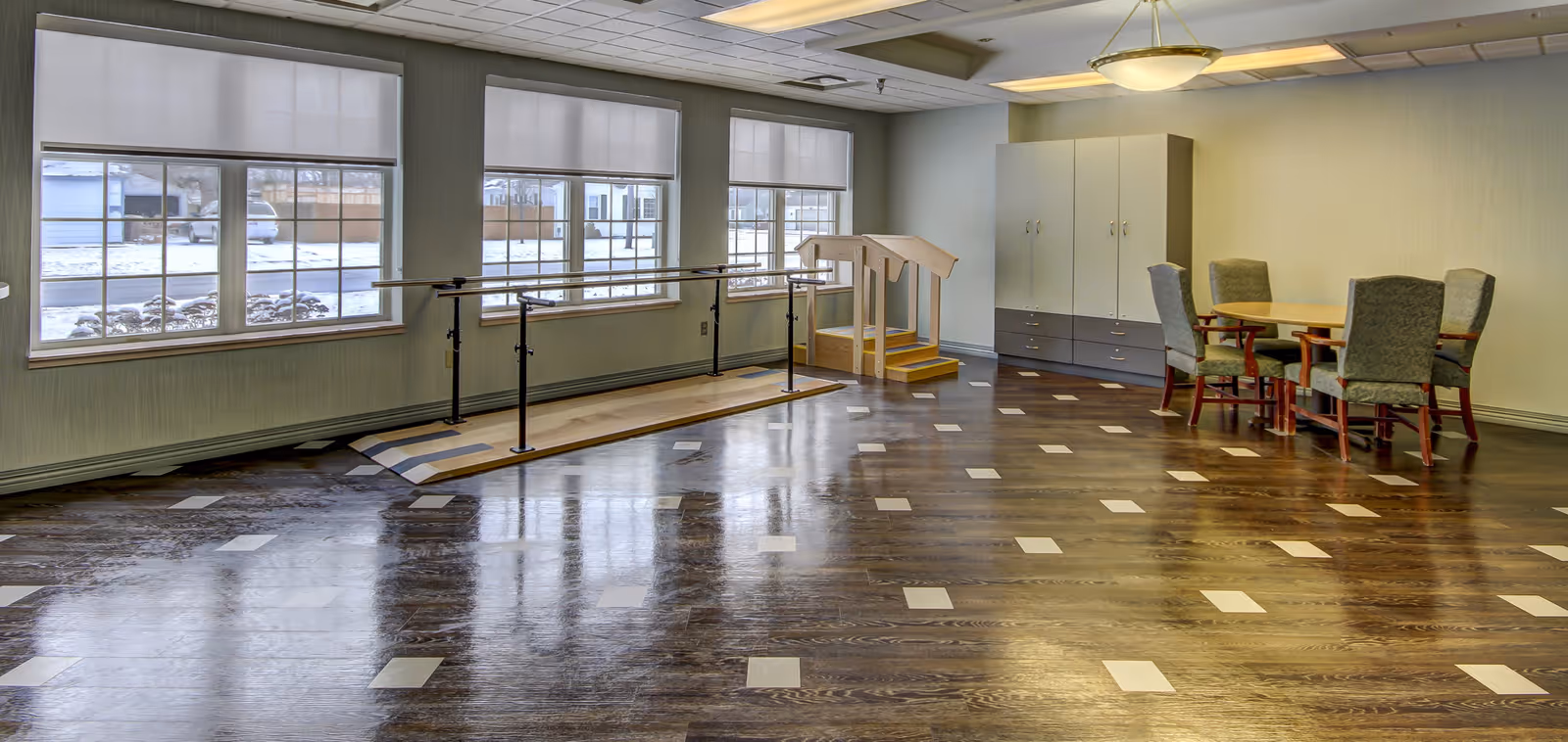 A spacious room with large windows showing a snowy outdoor scene. Inside, there is a wooden parallel bar setup for physical therapy or rehabilitation, a small set of stairs with handrails, a table with four upholstered chairs, and a large cabinet. The floor is dark wood with light square inlays, and the ceiling has recessed lighting and a hanging light fixture.