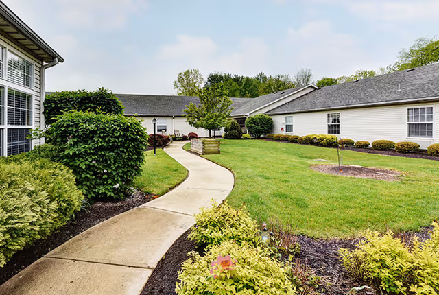 A curved concrete pathway winds through a well-maintained green lawn with various shrubs and small trees, surrounded by single-story white buildings with gray roofs under a partly cloudy sky.