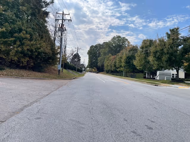 A wide paved road flanked by trees and utility poles on both sides under a partly cloudy sky during daytime.