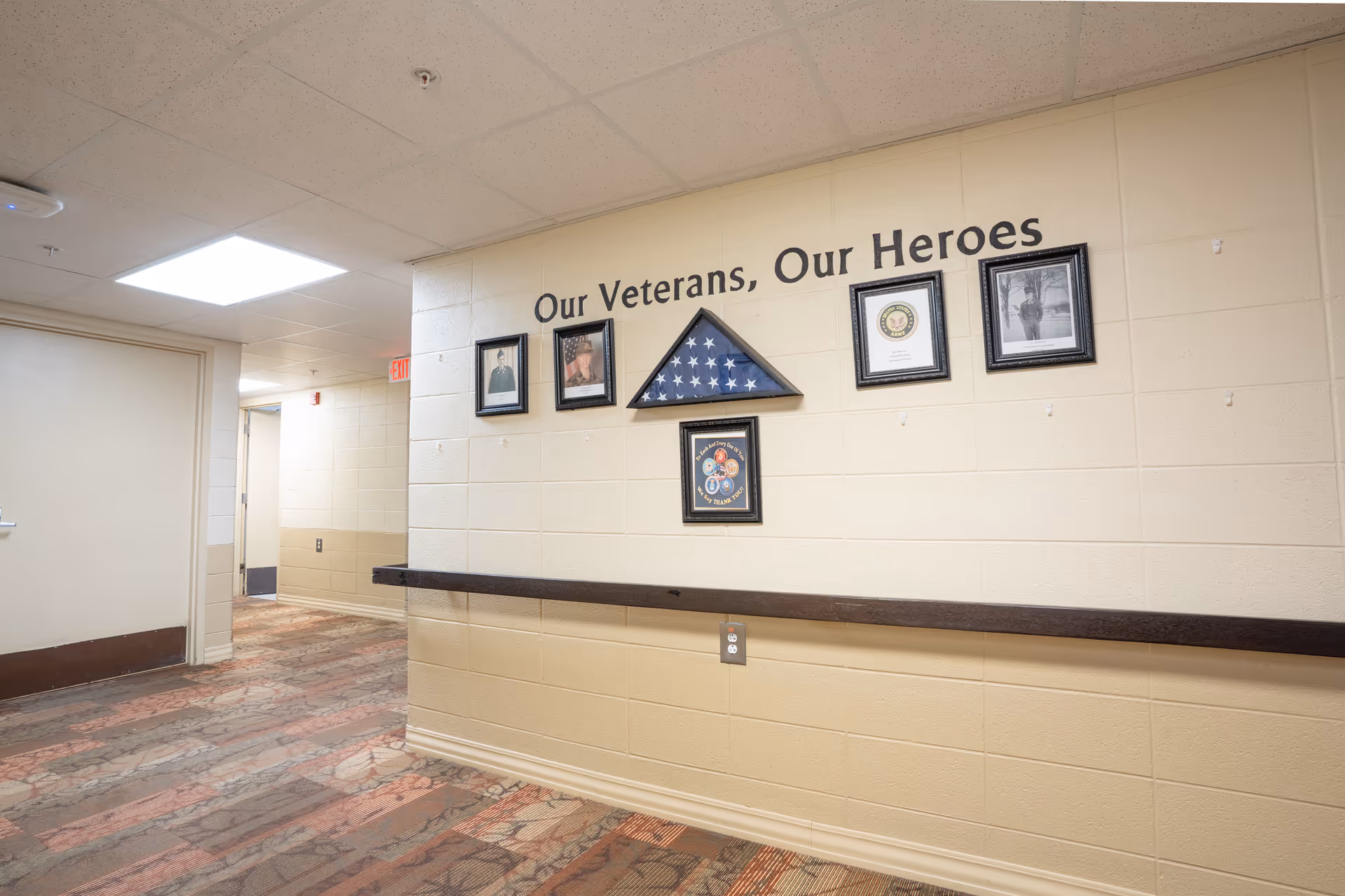 A hallway in a senior living facility with a display on the wall honoring veterans. The display includes framed photos, a folded American flag in a triangular case, and a plaque. Above the display, the text reads 'Our Veterans, Our Heroes'. The hallway has beige walls, a patterned carpet, and a handrail along the wall.