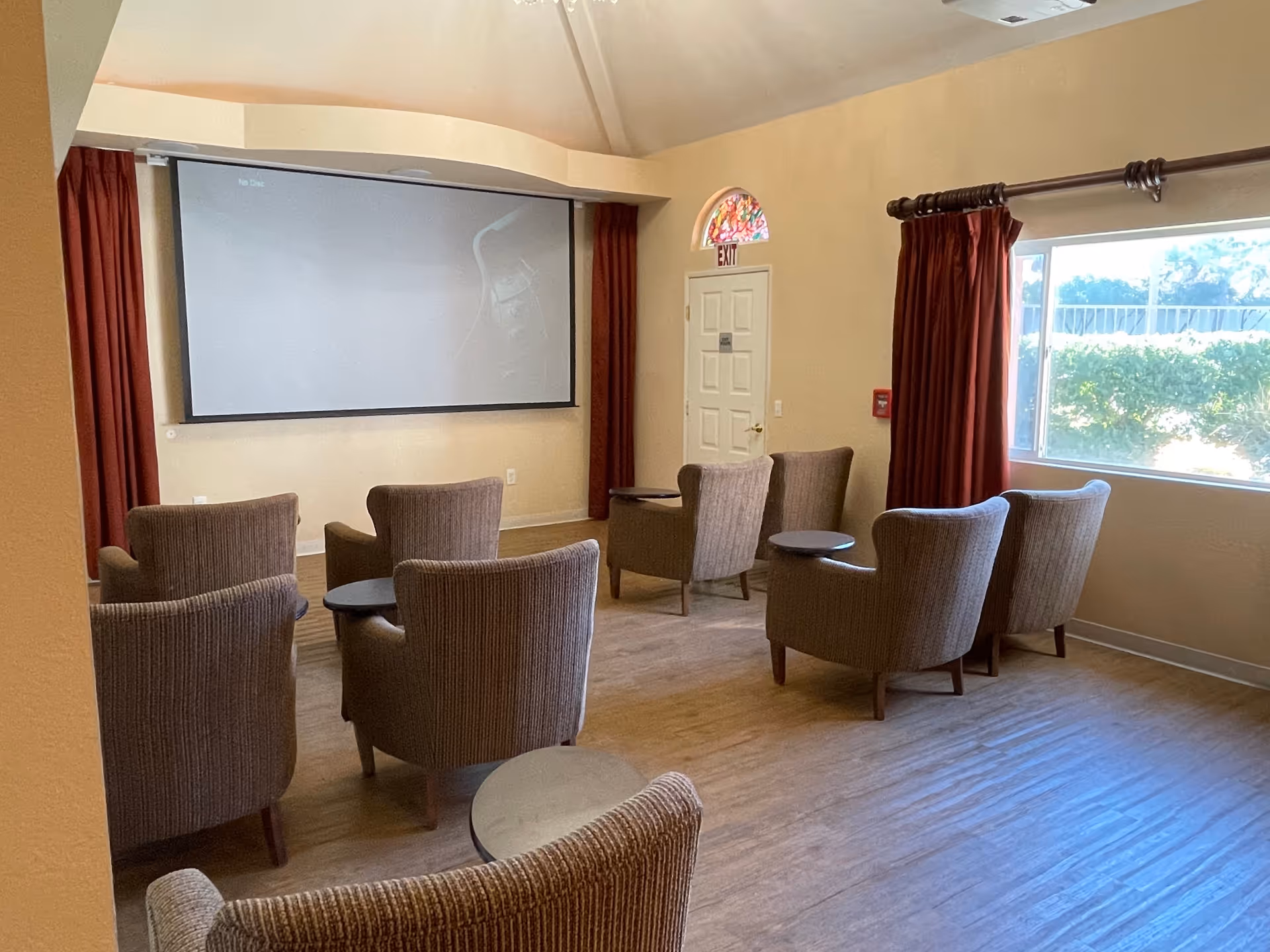 A small theater room with eight upholstered armchairs arranged in two rows facing a large projector screen. The room has beige walls, a window with red curtains on the right, and an exit door with a stained glass window above it at the back.