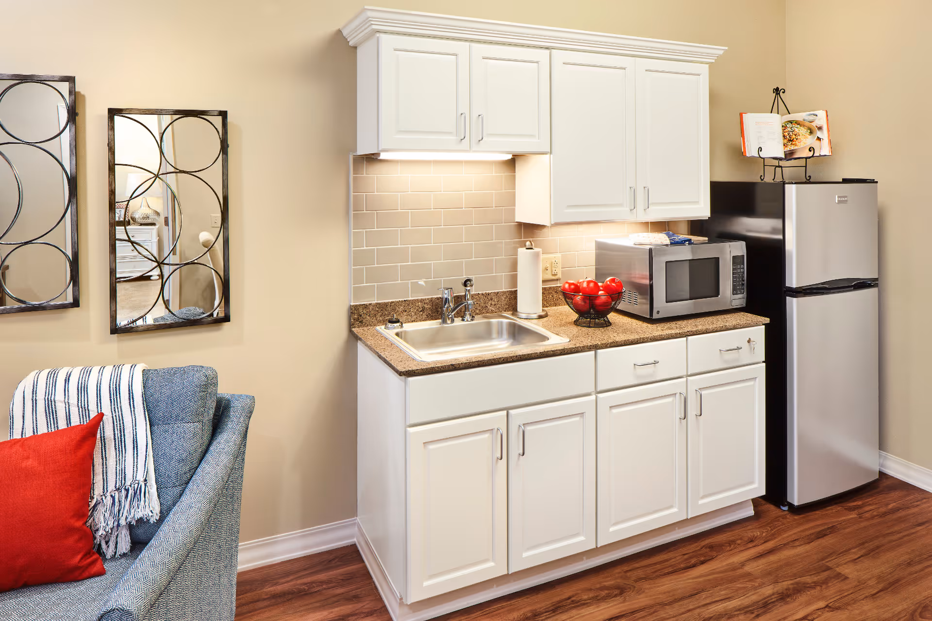 Small kitchen area with white cabinets, a stainless steel sink, a microwave, a bowl of red apples, and a stainless steel refrigerator. To the left, part of a blue upholstered chair with a red pillow and a striped throw blanket is visible. Two decorative mirrors with circular designs hang on the beige wall above the chair.