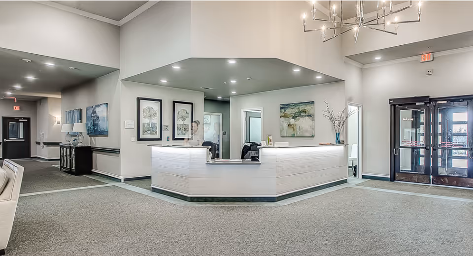 Bright, spacious reception lobby with a central front desk, seating area, artwork, and double glass entrance doors.