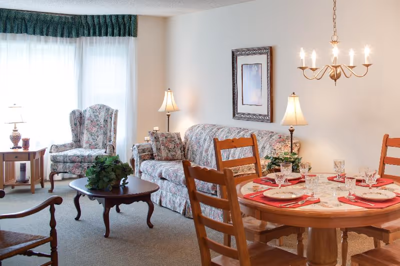 A cozy living and dining area with floral upholstered sofa and armchair, wooden coffee table with a plant, wooden dining table set with plates, glasses, and red napkins, two table lamps, a chandelier, and a framed picture on the wall.