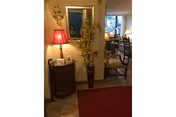 Interior view of a senior living facility showing a small wooden cabinet with a red lamp and decorative items on top, a tall vase with yellow flowers beside it, a mirror on the wall, and a dining area with chairs and a table visible in the background.