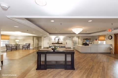 A spacious and well-lit senior living facility lobby with a reception desk on the right, a seating area with chairs and a table on the left, and a decorative table with flowers in the center. The room features wood flooring, neutral-colored walls, ceiling lights, and a large chandelier.