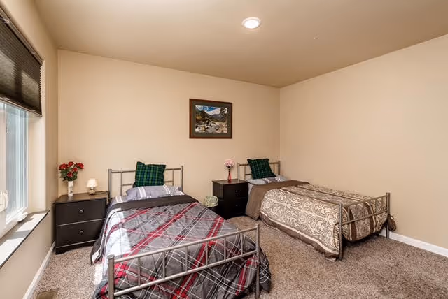 A simple bedroom with two single metal frame beds, each with different patterned bedding. Between the beds is a black nightstand with a small pink flower vase and a green decorative item. There is a framed landscape picture on the beige wall above the nightstand. A window with a black blind is on the left side of the room, and the floor is carpeted in a neutral tone.