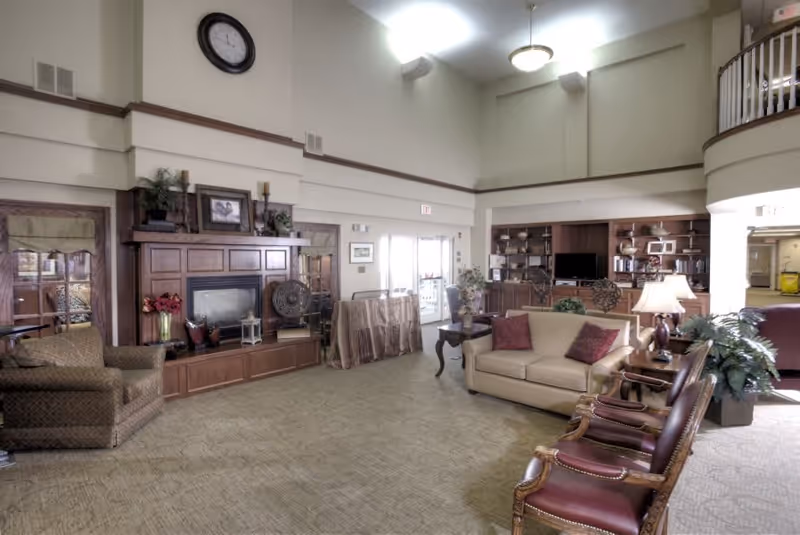A spacious living room area in a senior living facility with high ceilings and soft lighting. The room features a beige sofa with two red pillows, several wooden chairs with leather seats, a wooden coffee table with a lamp, and a large wooden entertainment center with a TV and shelves filled with books and decorative items. There is a fireplace with a wooden mantel adorned with plants, candles, and framed pictures. The room has carpeted flooring and large windows allowing natural light to enter.