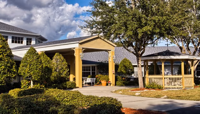 Exterior view of a senior living facility named Heron House, featuring a covered entrance with columns, well-maintained landscaping with bushes and trees, and a wooden gazebo on the right side under a partly cloudy sky.
