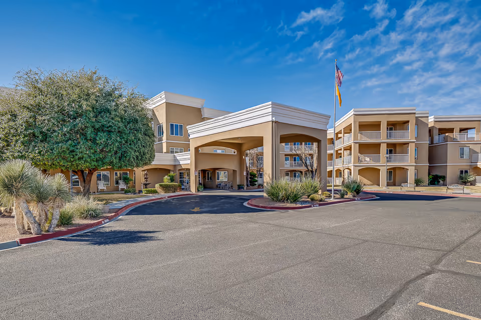 Entrance and driveway of a multi-story beige senior living building with balconies, landscaping, and flagpoles under a blue sky.