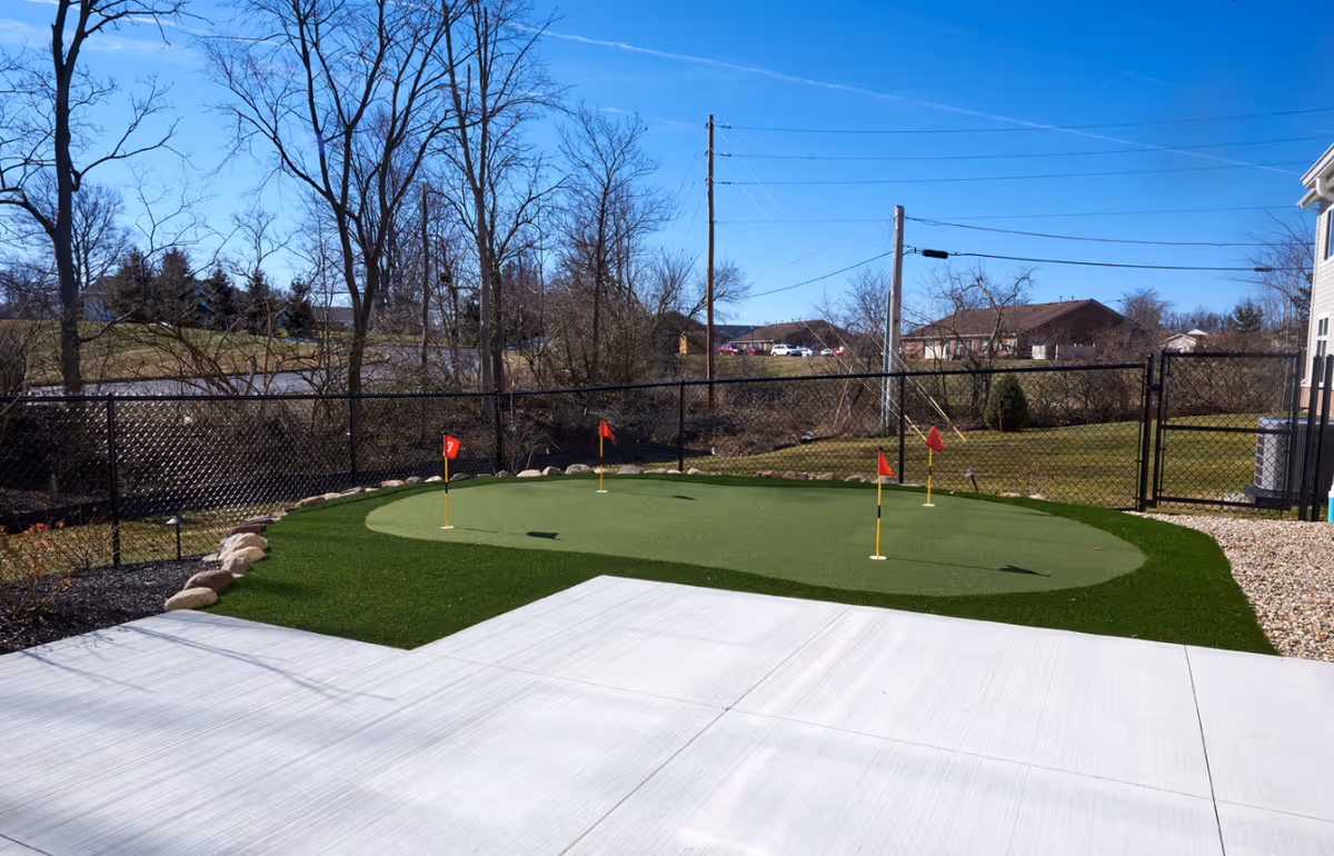Outdoor putting green with five small red flags on a sunny day, surrounded by a black chain-link fence and some leafless trees in the background.
