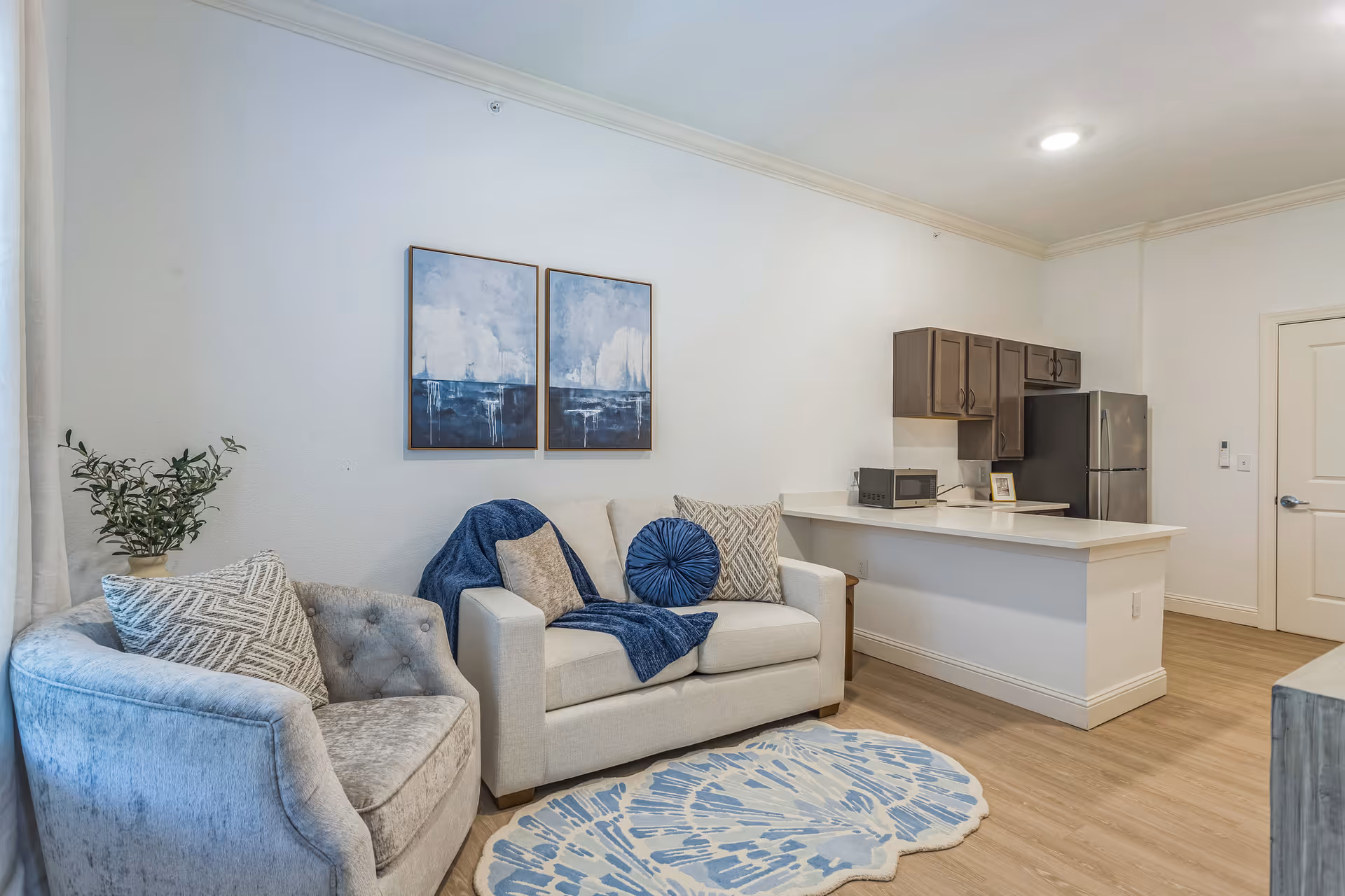A cozy living area featuring a light gray armchair and a white loveseat adorned with blue and beige pillows and a blue throw blanket. Above the loveseat are two framed abstract paintings in blue and white tones. The room has light wood flooring and a small kitchen area with dark wood cabinets, a microwave, and a stainless steel refrigerator in the background. A blue and white patterned rug is placed on the floor in front of the seating.