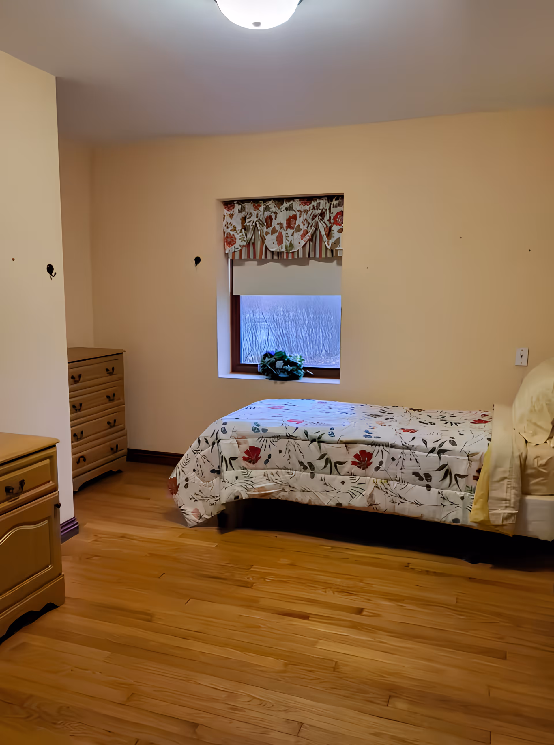 Simple single bedroom with a twin bed in floral bedding, wooden dressers, and a small window with a floral valance.