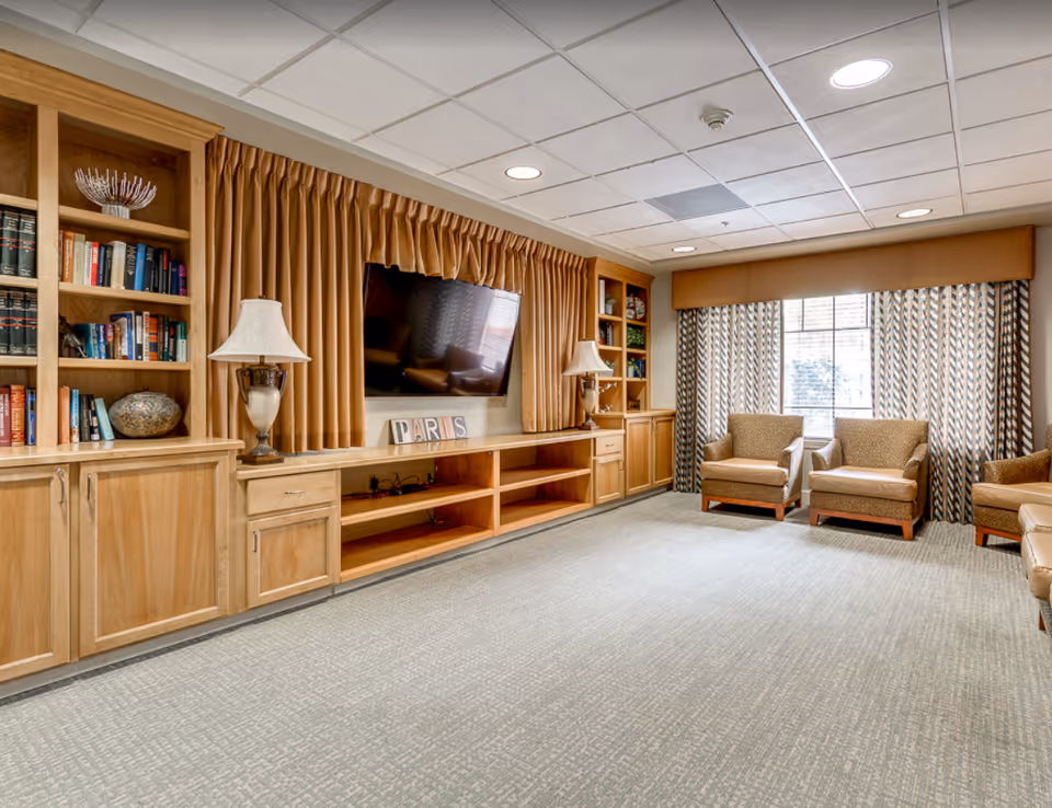 A cozy living room area with built-in wooden shelves and cabinets, a large flat-screen TV mounted on the wall with brown curtains behind it, two table lamps on either side of the TV, three upholstered armchairs near a window with patterned curtains, and a light-colored carpeted floor.