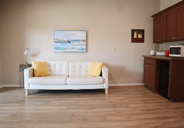 A white sofa with two yellow pillows sits against a beige wall with framed artwork, hardwood floors, and a small kitchenette to the right.
