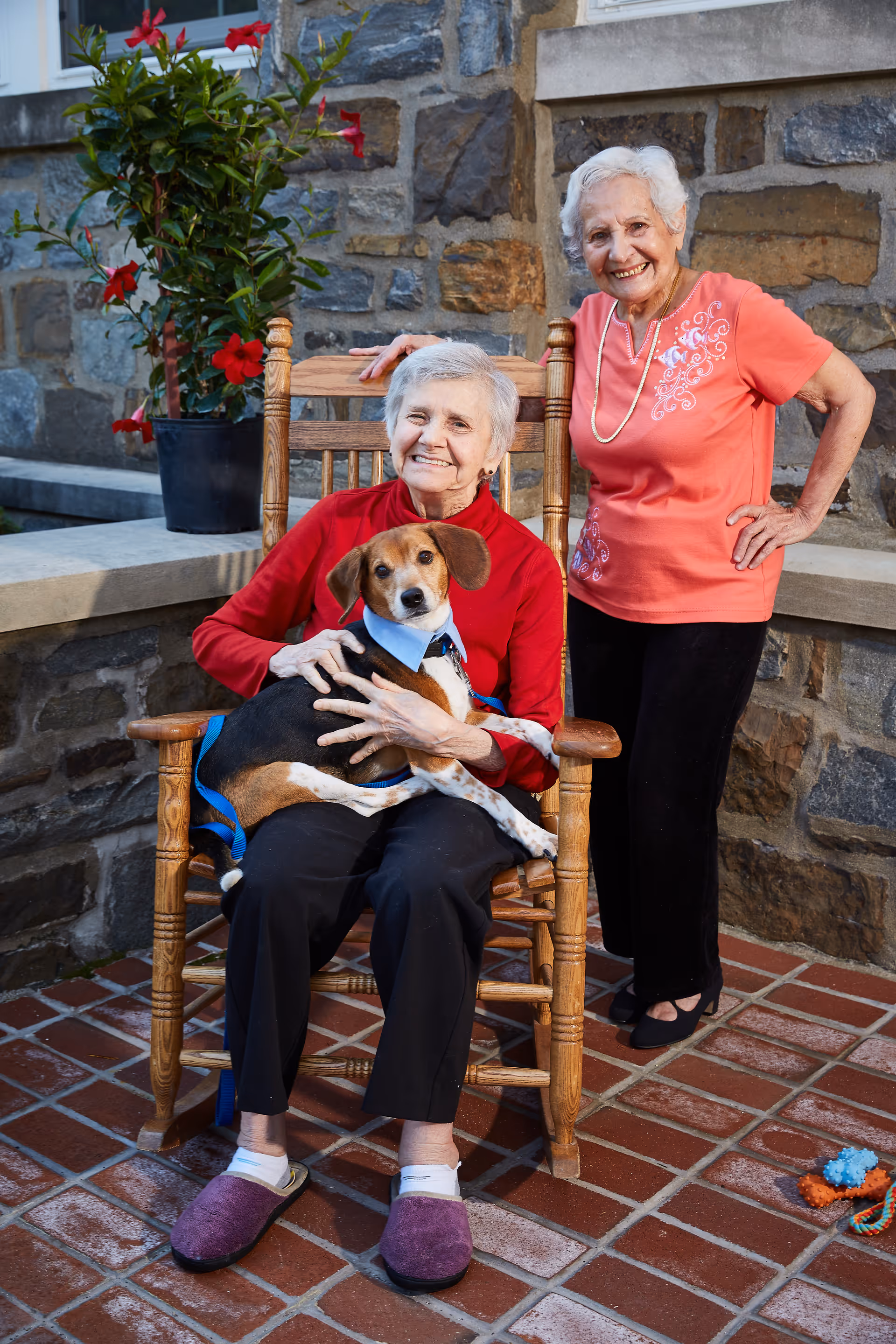 Two elderly women smiling outdoors in front of a stone wall. One woman is sitting in a wooden rocking chair holding a dog wearing a blue collar and leash. The other woman is standing beside her with one hand on her hip. There is a potted plant with red flowers behind them and dog toys on the tiled ground.