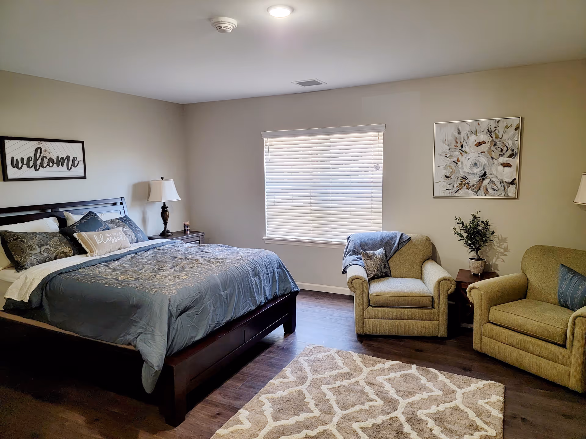 A cozy bedroom with a large bed featuring a dark wooden frame and blue patterned bedding. There are two beige armchairs with decorative pillows and a small wooden side table with a potted plant between them. A window with closed blinds is centered on the wall, and the walls are decorated with a 'welcome' sign and a floral painting. The floor is dark wood with a beige patterned rug.