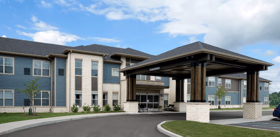 Exterior view of a senior living facility named Harmony at White Oaks, showing a large covered entrance with wooden pillars and a two-story building with blue and white brick facade under a partly cloudy sky.