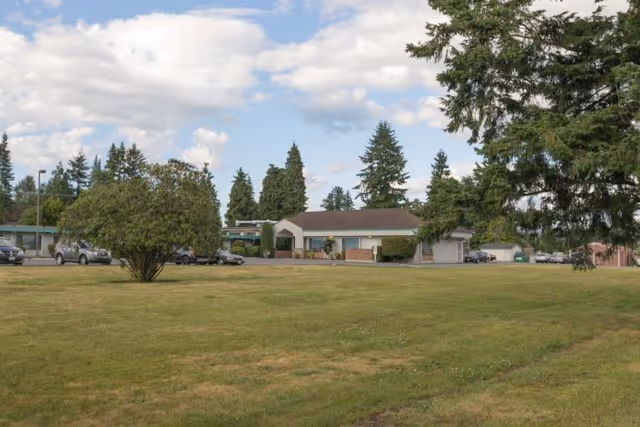 A wide grassy lawn with a few scattered trees and bushes in front of a single-story building with a brown roof, surrounded by parked cars and tall evergreen trees under a partly cloudy sky.