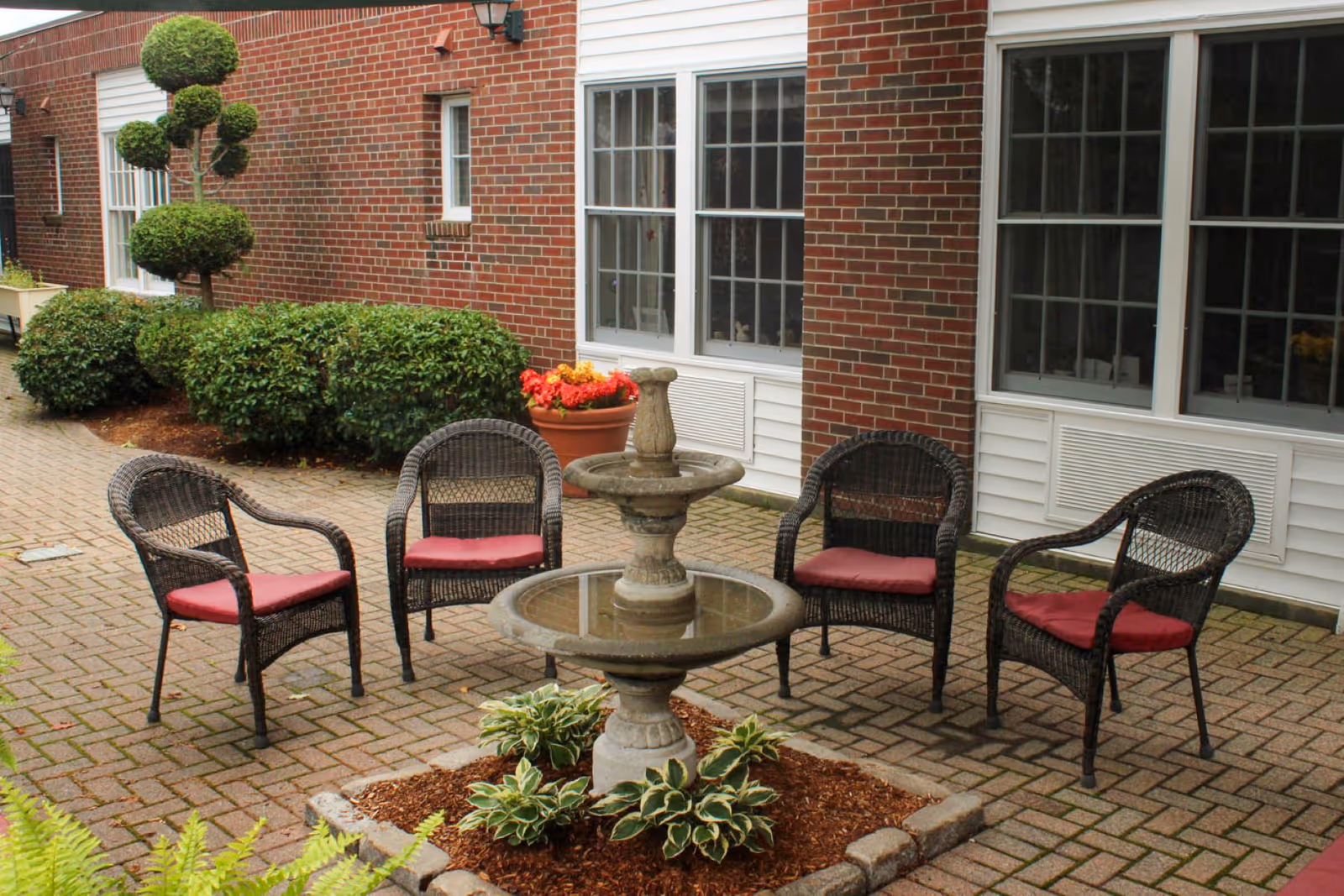 Outdoor patio area with four wicker chairs with red cushions arranged around a two-tier stone fountain. The patio is paved with bricks and bordered by neatly trimmed bushes and a topiary tree. The background shows a brick building with white-framed windows and a flower pot with red flowers.