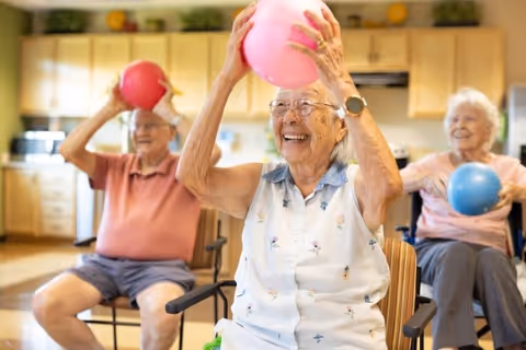 Three elderly individuals sitting on chairs in a bright kitchen area, each holding a colorful exercise ball above their heads and smiling during a group activity.