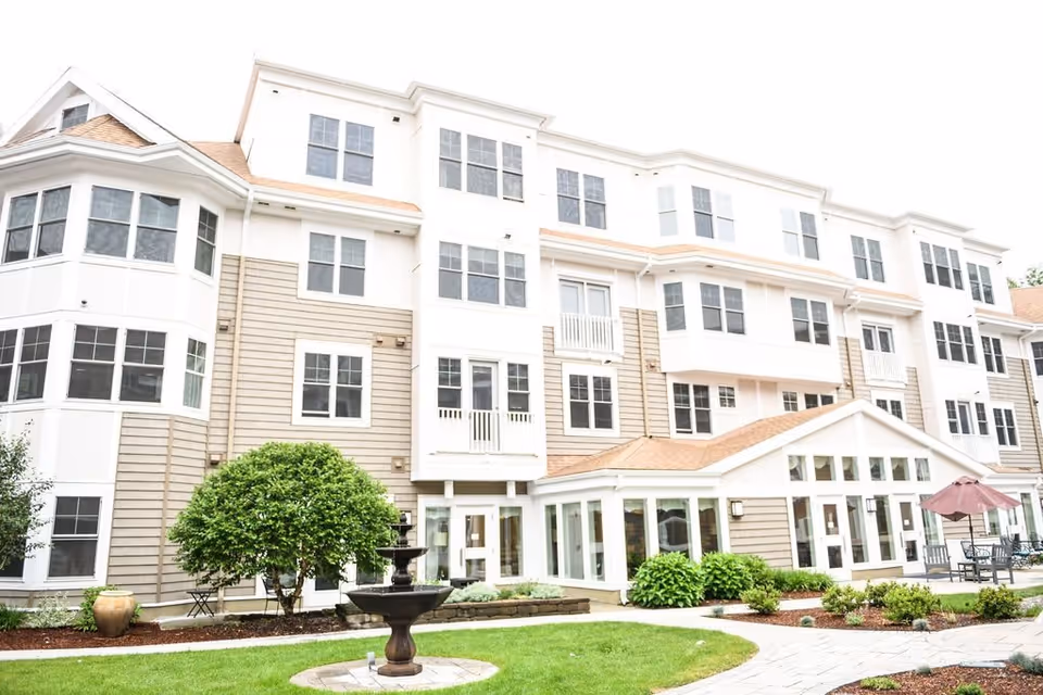 Exterior view of a multi-story assisted living facility with beige and white siding, numerous windows, a small fountain, green shrubs, and a patio area with tables and umbrellas.