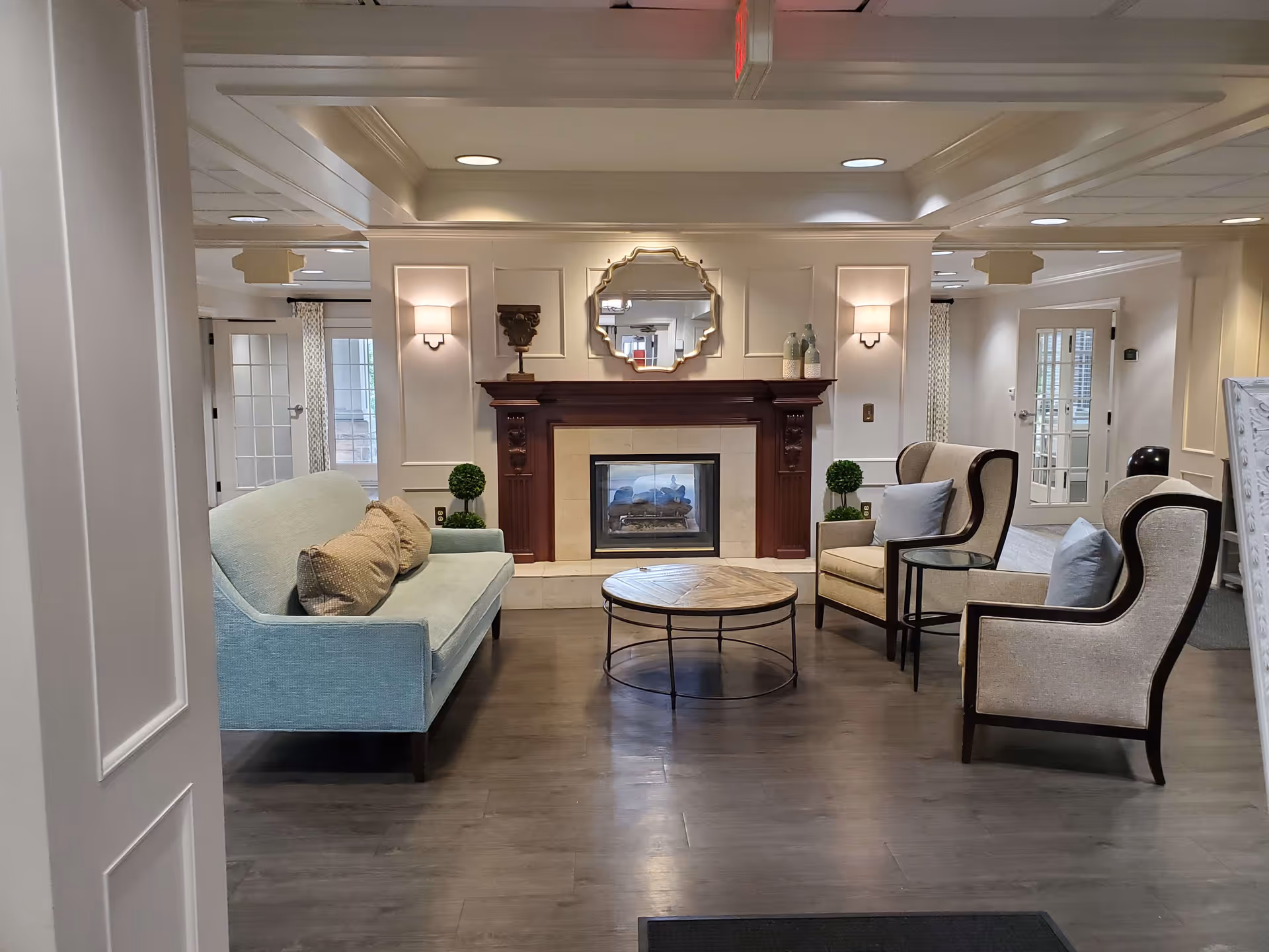 A cozy living room area in Forest Hills Place featuring a light blue sofa with beige pillows, two beige armchairs with light blue pillows, a round wooden coffee table, and a fireplace with a decorative mirror above it. The room has soft lighting, white walls with paneling, and glass doors in the background.